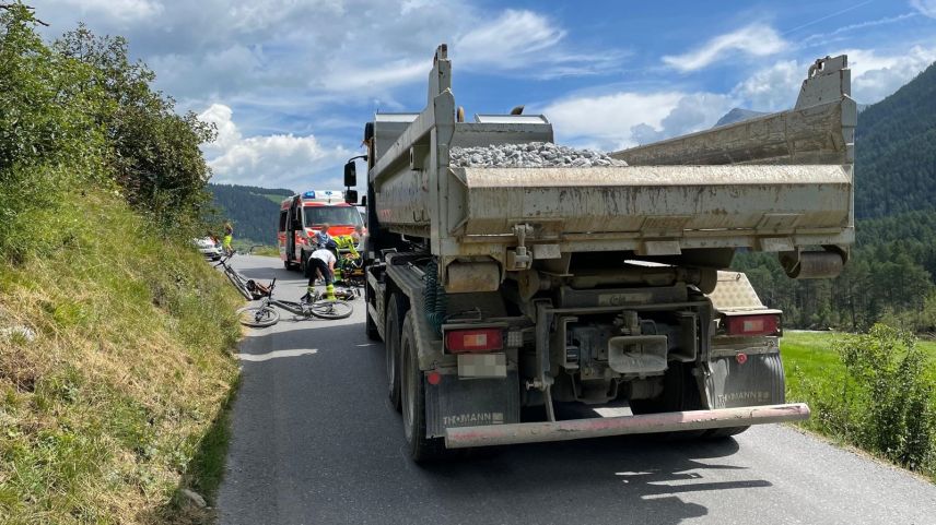 Der Lastwagen von hinten. Vor diesem ein Fahrrad am Boden und sowie Rettungskräfte und eine Ambulanz (Foto: Kantonspolizei Graubünden).