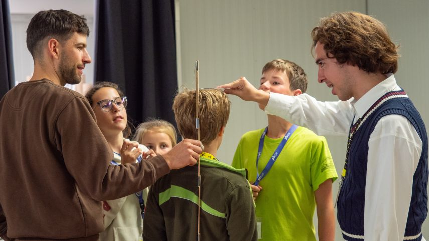 Gildo Hohenegger von der Schreinerei Pichalain SA in Fuldera (links) und PH-Student Marcel von Salis machen mit den Kindern eine Lockerungsübung bei der die Kinder ihre Grösse schätzen müssen. Danach wird gemeinsam nachgemessen.