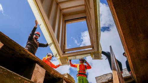 Flughelfer der Heli Bernina und Mitarbeiter der Holzbaufirma Nusser manövrieren das «heranschwebende» Element des neuen Dachausstiegs punktgenau auf die vorbereitete Dachöffnung. Foto: Jon Duschletta